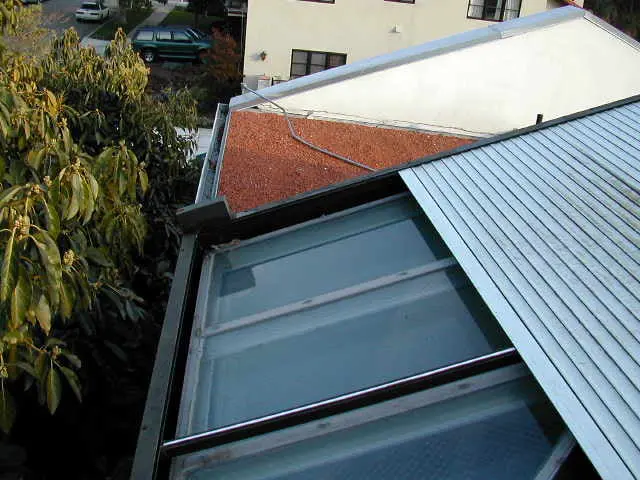 Aerial view of two roof sections, one with a gravel surface and the other with glass panels, surrounded by trees and a glimpse of parked cars in the background.