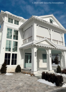 A modern white house with large windows and a covered porch, featuring a sloped roof and decorative trim. Snow is visible on the ground and around the landscaping, creating a winter scene.